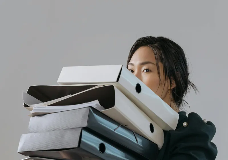 Person overwhelmed carrying a stack of disorganized files and binders